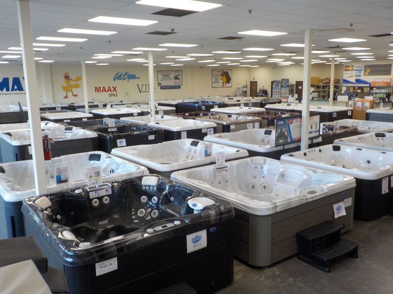 A showroom floor filled with rows of various hot tubs and spas under a drop ceiling with many recessed lights.