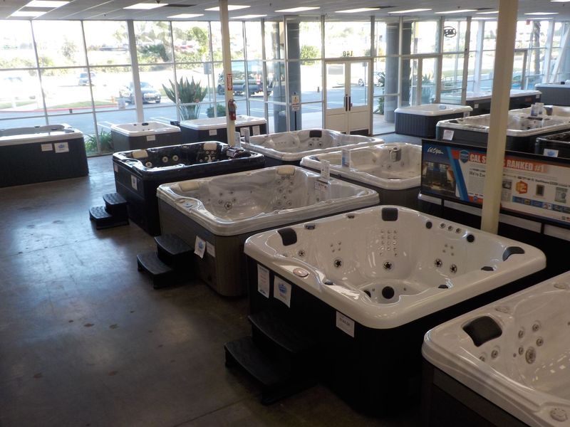 A showroom filled with various hot tubs, featuring white and dark grey models arranged on a concrete floor by large windows.