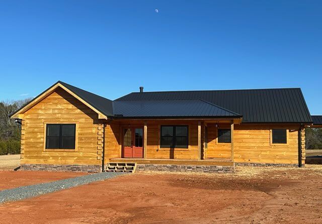 Wooden cabin with black roof and porch on a red dirt lot under a clear blue sky