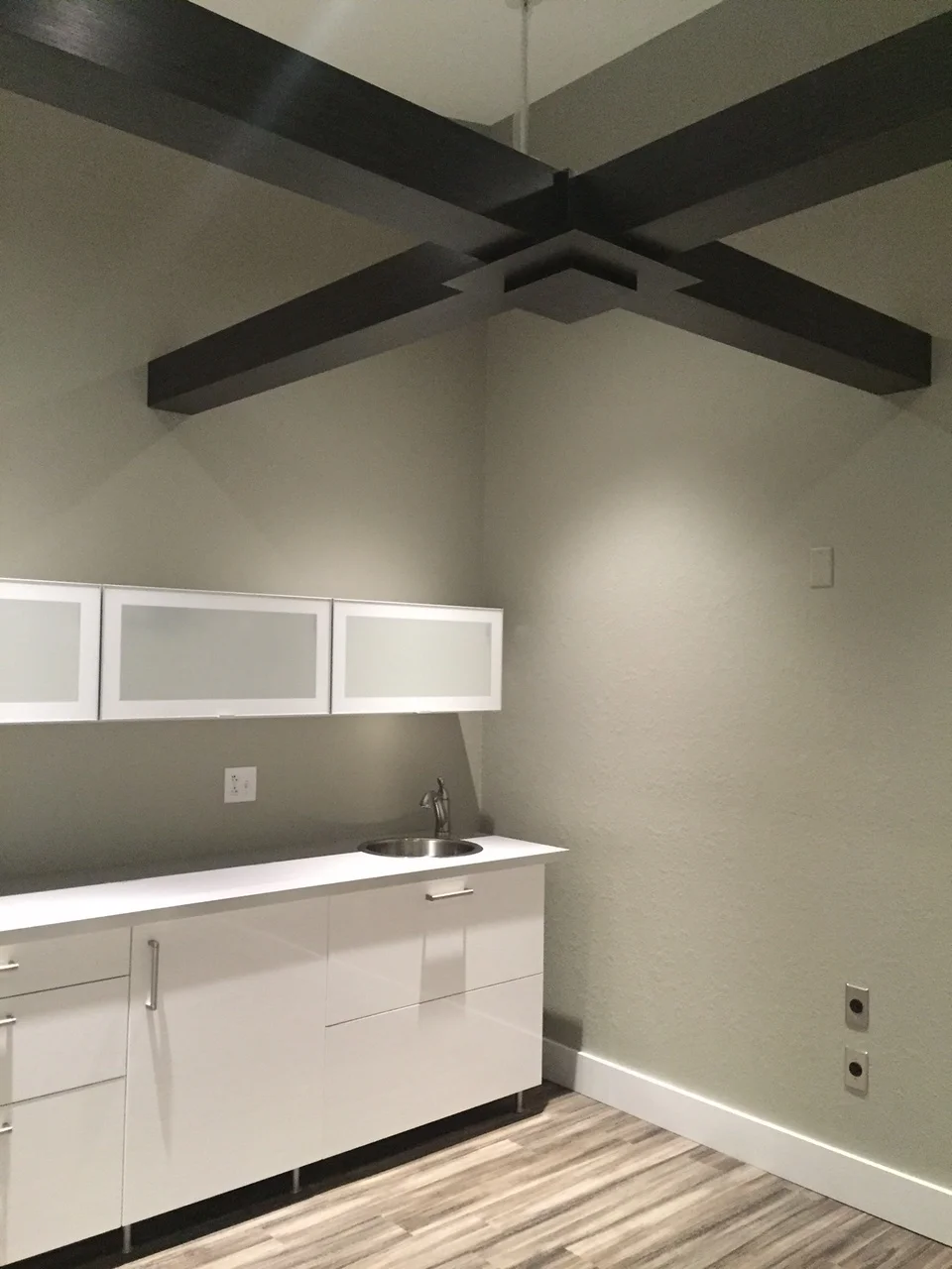 A modern, minimalist bathroom with white cabinets, sink, and wooden-like flooring; dark beams cross the ceiling.