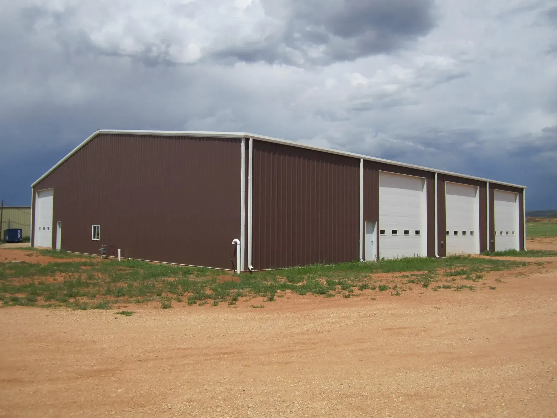 Brown metal building with white doors and overhead garage doors, in a field under cloudy sky.