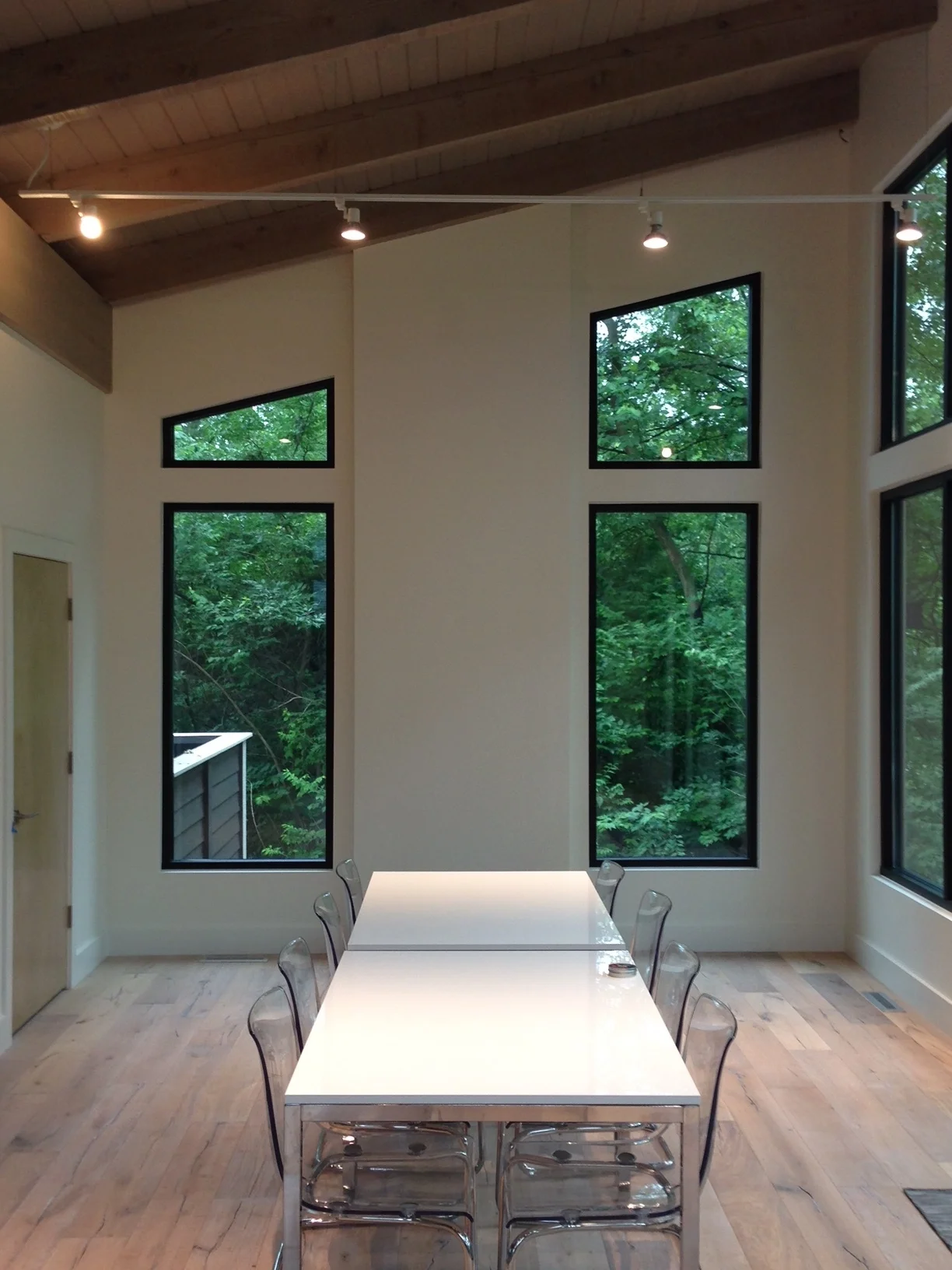 Modern dining room with white table, clear chairs, and tall windows overlooking trees.