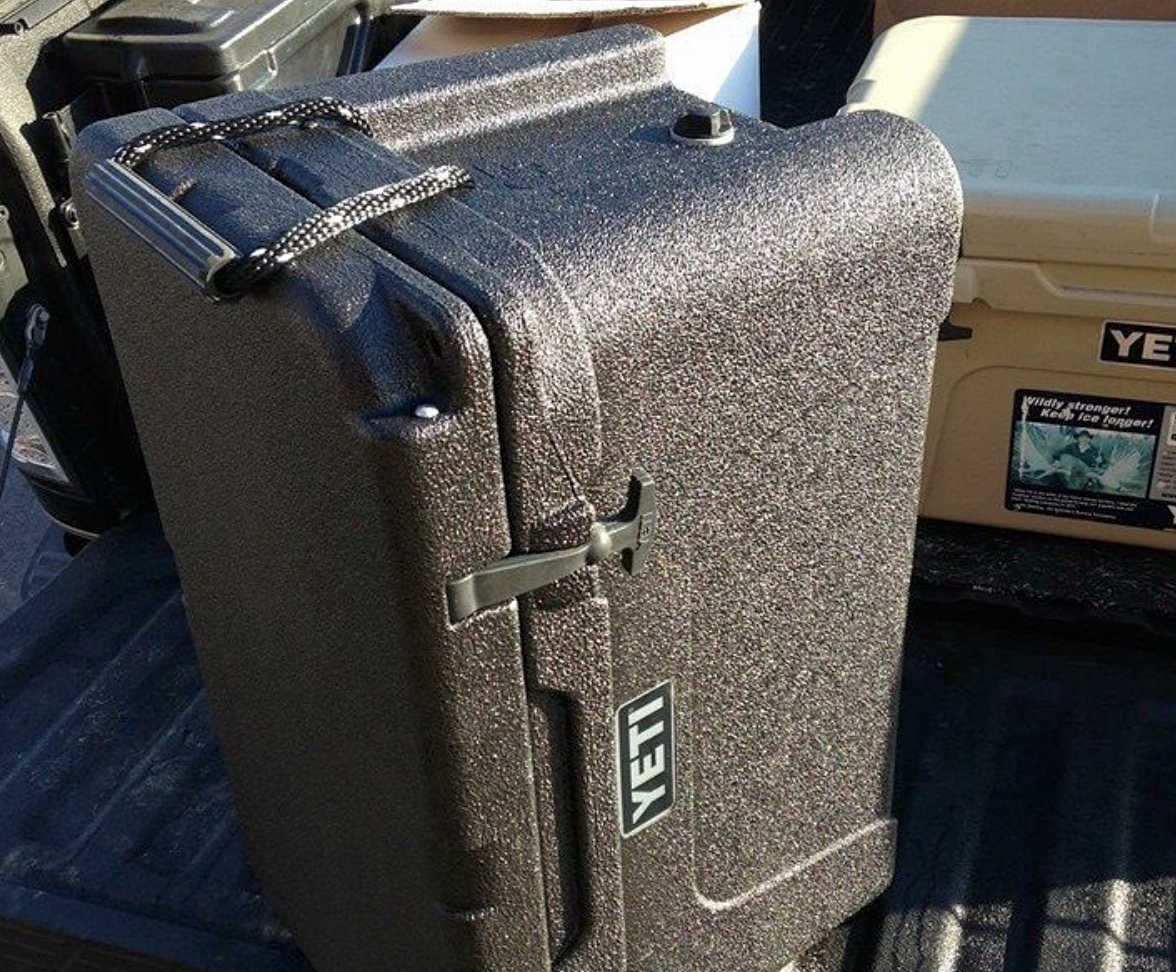 A textured, dark-colored Yeti cooler with a handle and latch sits on a truck bed.