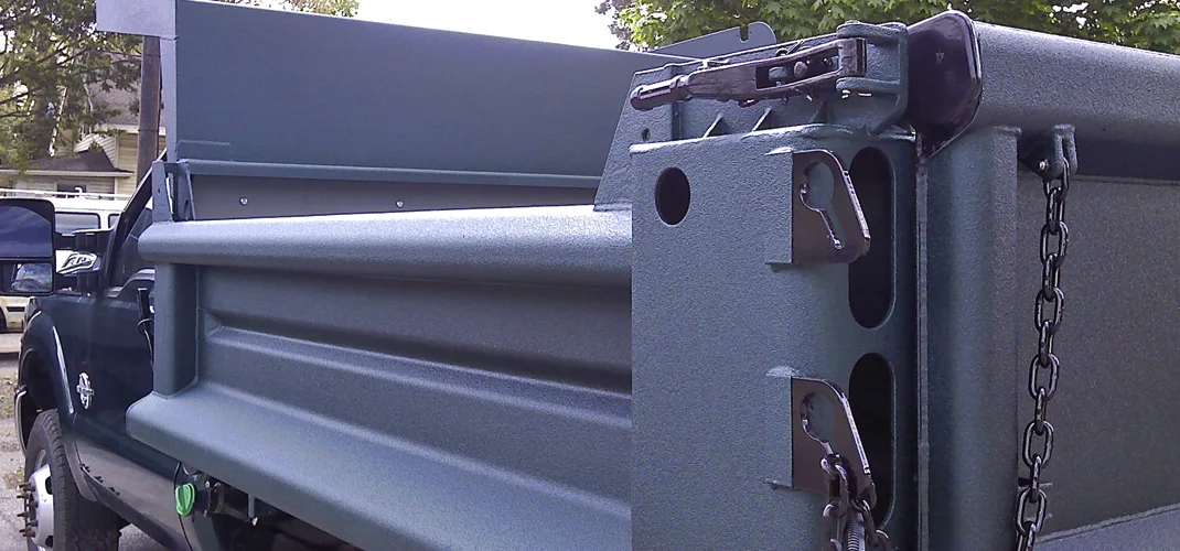 A close-up of a dark green dump truck bed, showing the details of the hinges, chains, and rounded bar.