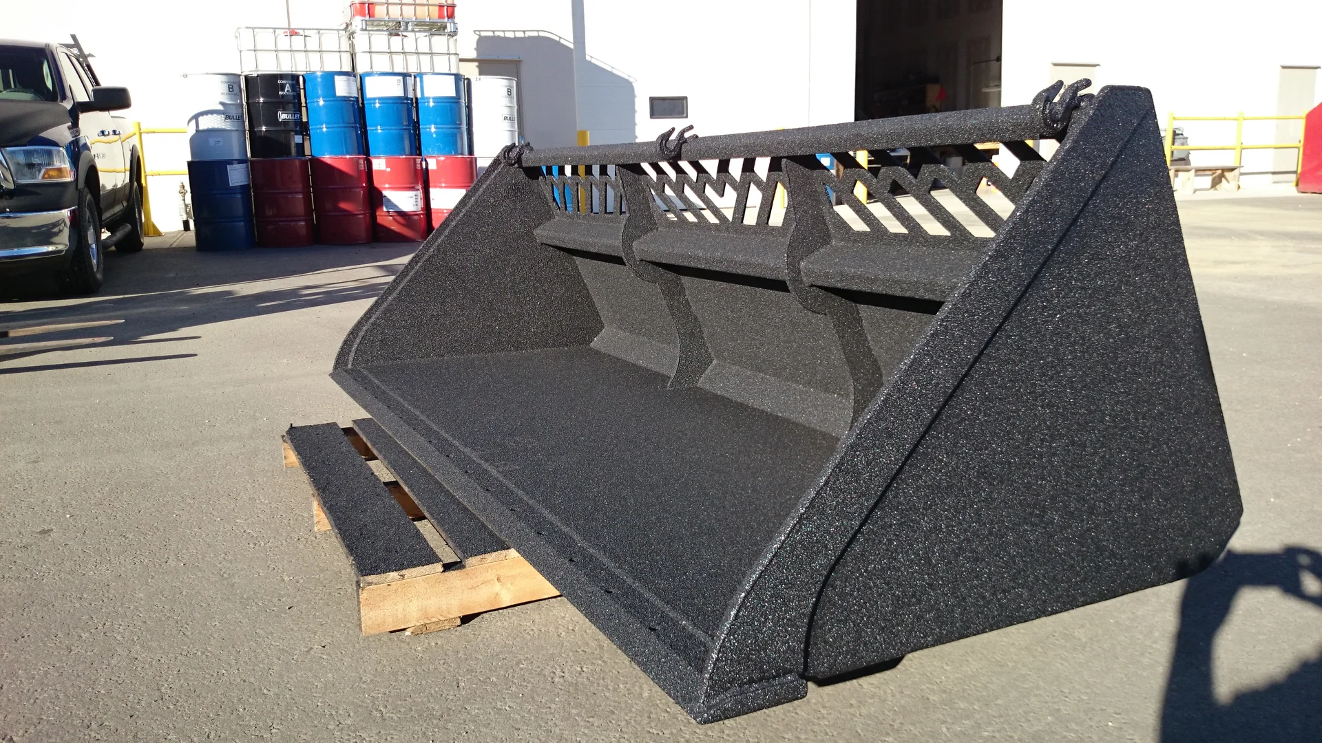 Black front-end loader bucket on a wooden pallet outdoors. In the background are barrels and a building.