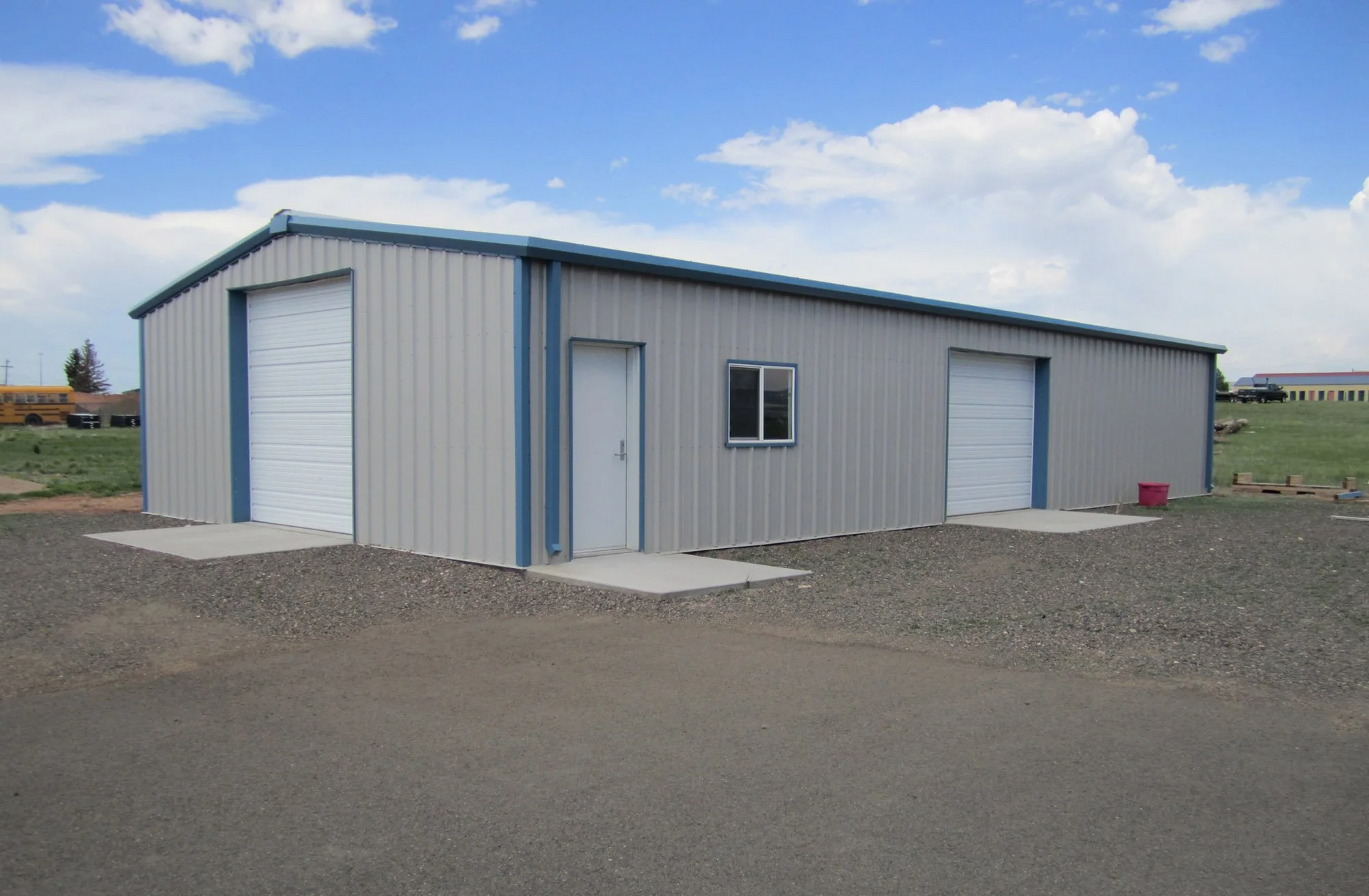 Gray metal building with two garage doors, a door, and a window; gravel lot, blue sky.