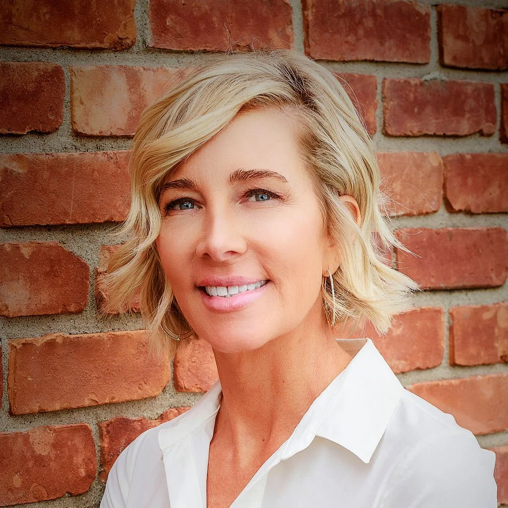 Blonde woman with short hair smiles, wearing a white shirt, against a brick wall.