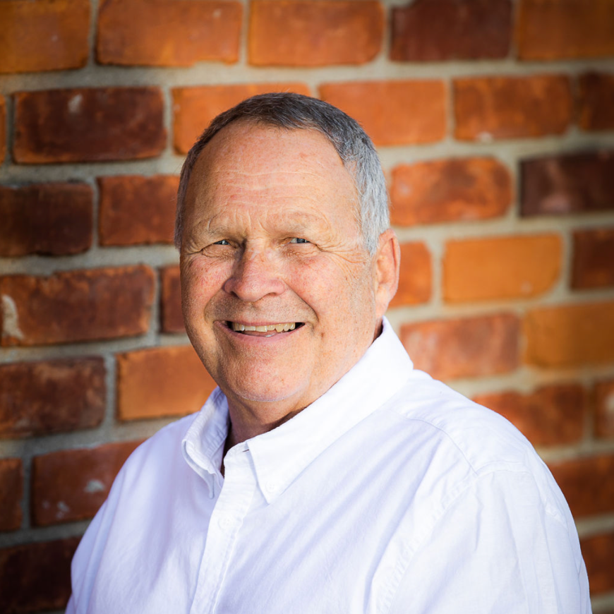 Man with gray hair smiles, wearing a white shirt against a brick wall background.