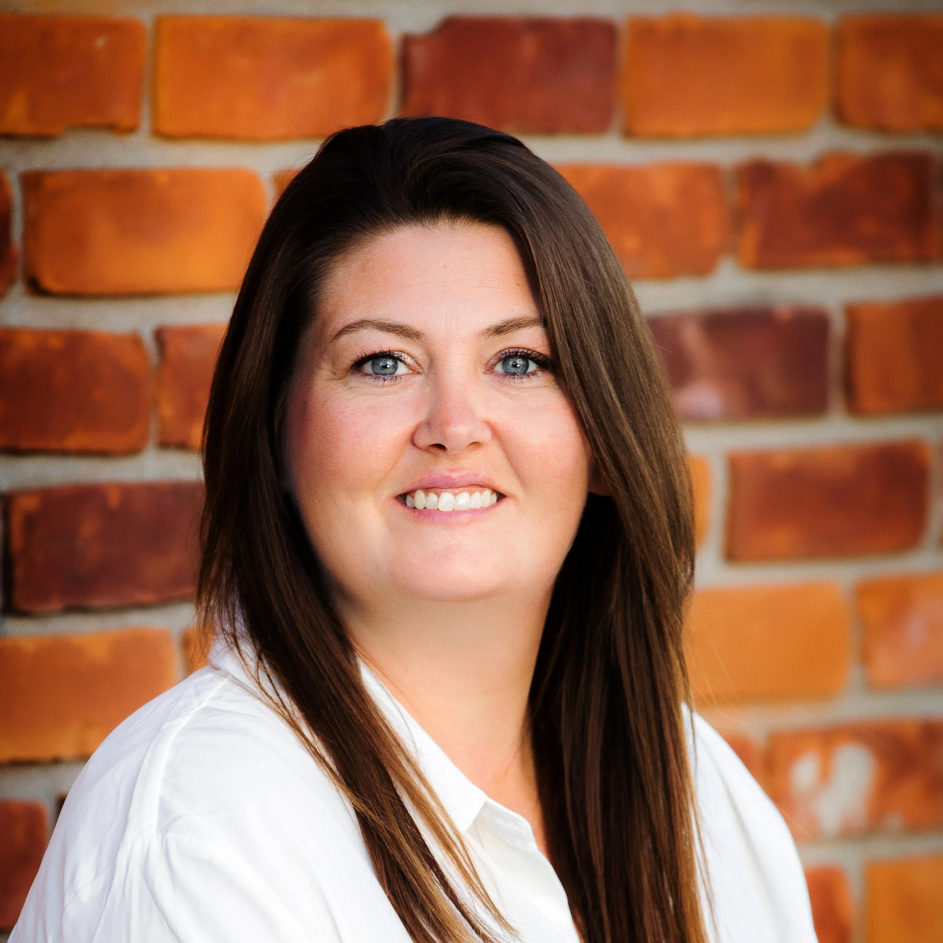 Woman with long brown hair, blue eyes, smiles, in front of a brick wall.