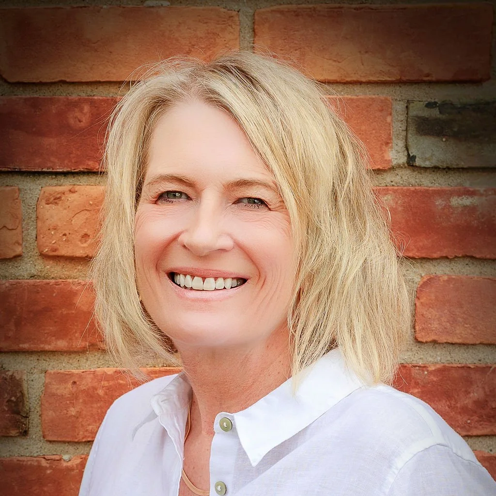 Blonde woman smiling, wearing a white shirt, against a brick wall.