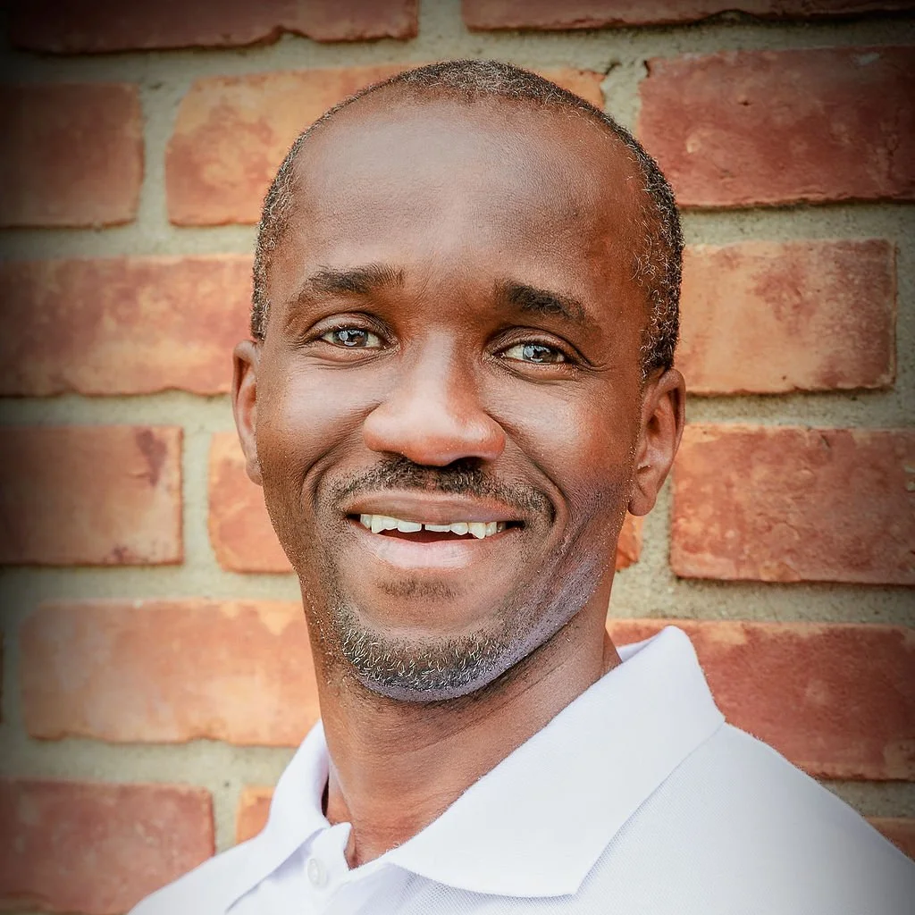 Smiling Black man in white shirt against a brick wall background.