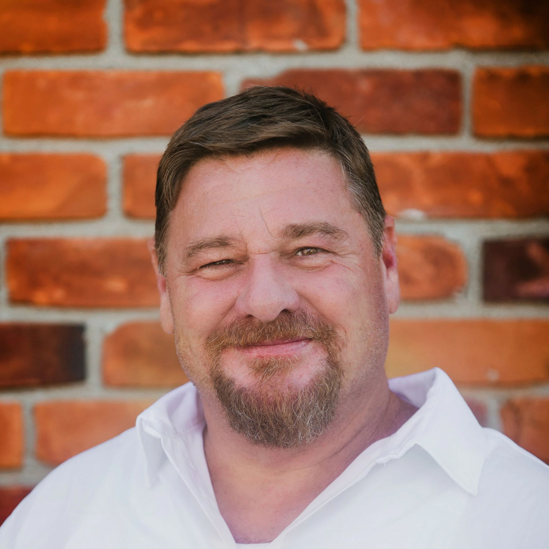 Man with a beard smiles in front of a brick wall. He wears a white collared shirt.