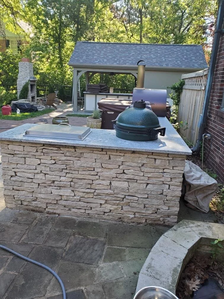 Outdoor stone-covered kitchen counter with grill, smoker, and sink in backyard setting.