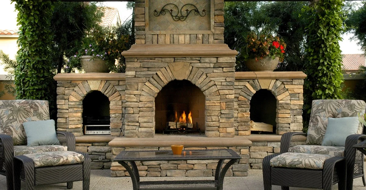 Stone outdoor fireplace with chairs and a coffee table, set in a backyard.
