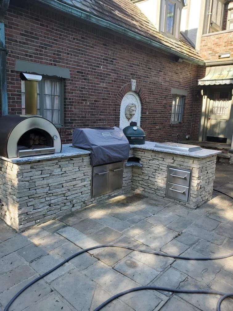 Outdoor kitchen with brick facade and stone countertop; includes grill, pizza oven, and Big Green Egg.