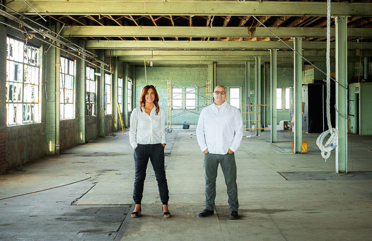 Two people stand in an empty, run-down warehouse. Woman in white shirt and black pants, man in white shirt and gray pants.