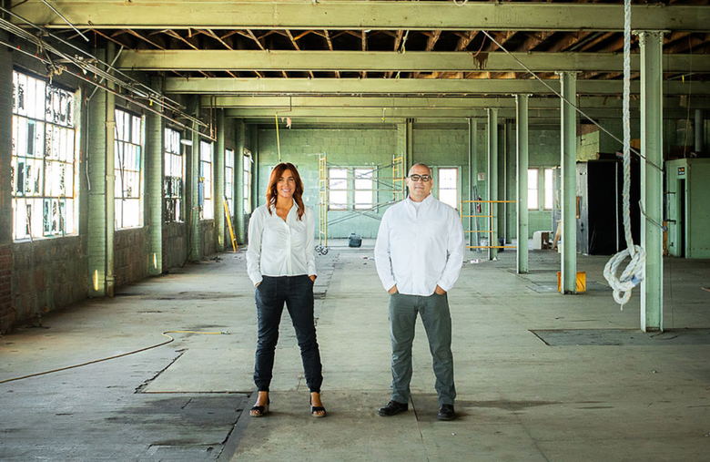 Two people stand in an empty warehouse. The woman wears a white shirt and black pants. The man wears a white shirt and gray pants.