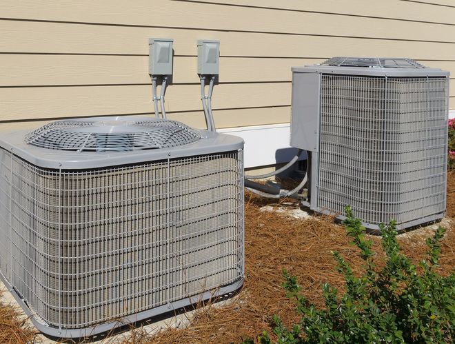 Two outdoor air conditioning units beside a beige house wall on a mulch bed