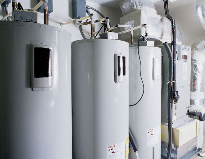Row of large gray industrial water heaters with pipes and wall-mounted controls in a utility room