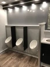 Three-stall gray restroom urinal area with white urinals and overhead lights, wood-look floor