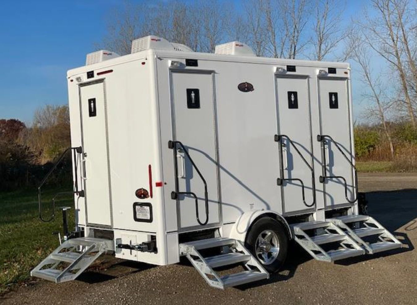 Portable white restroom trailer with multiple doors and steps parked outdoors on a paved lot.