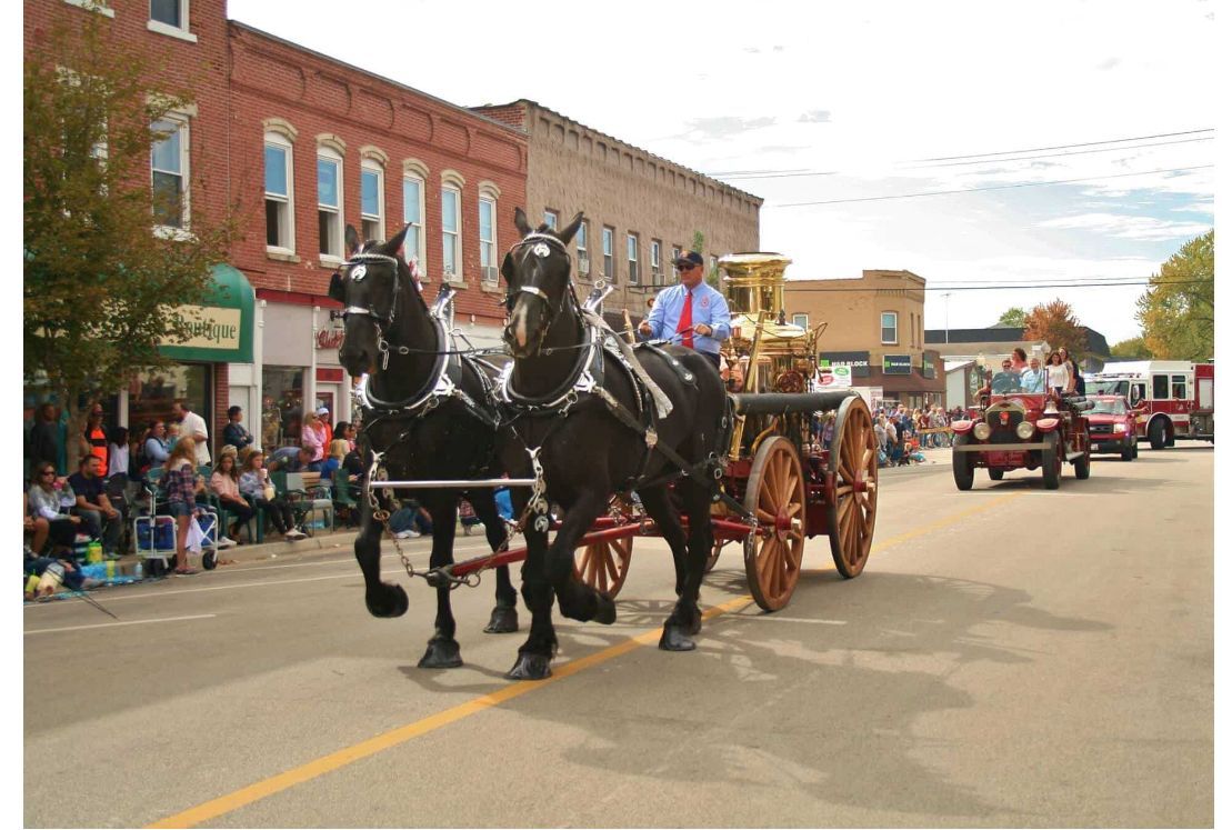 Horse-drawn carriage parade on a small-town street with spectators lining the sidewalks