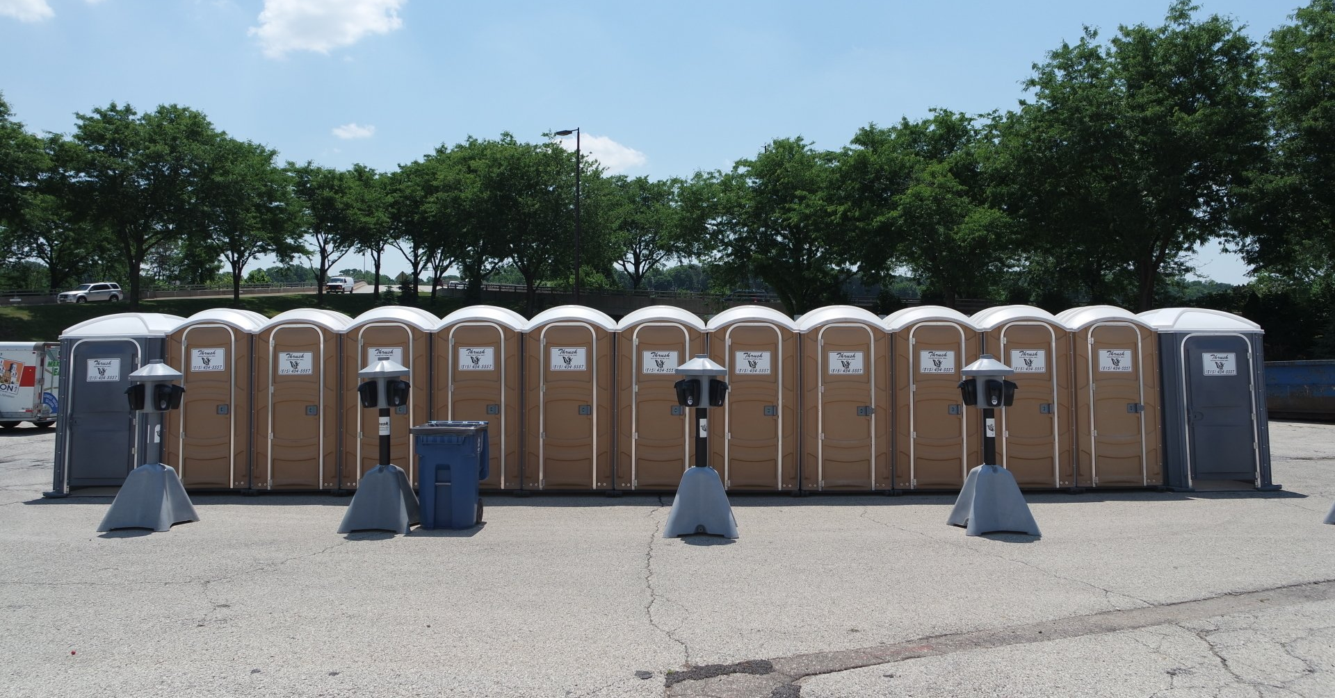 Row of tan and gray portable toilets behind concrete barriers in a parking lot under trees