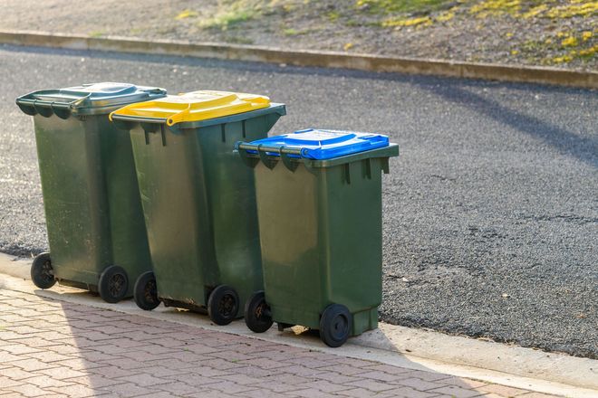Four wheeled curbside trash bins with green, yellow, and blue lids lined up on a street curb