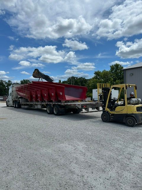 Yellow forklift unloading a red trailer truck in a gravel lot under a cloudy blue sky