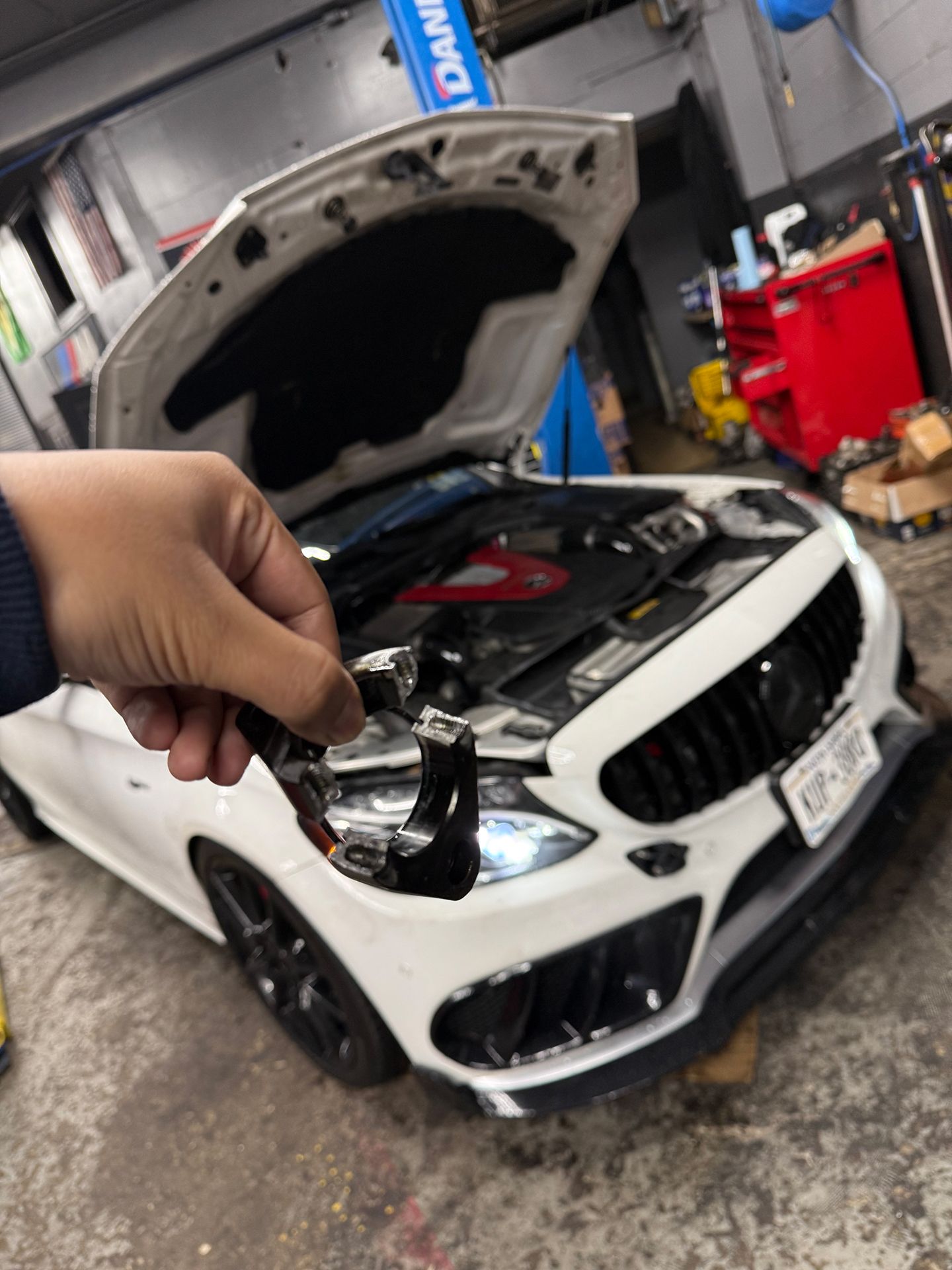Hand holding a broken part in front of a white car with the hood open