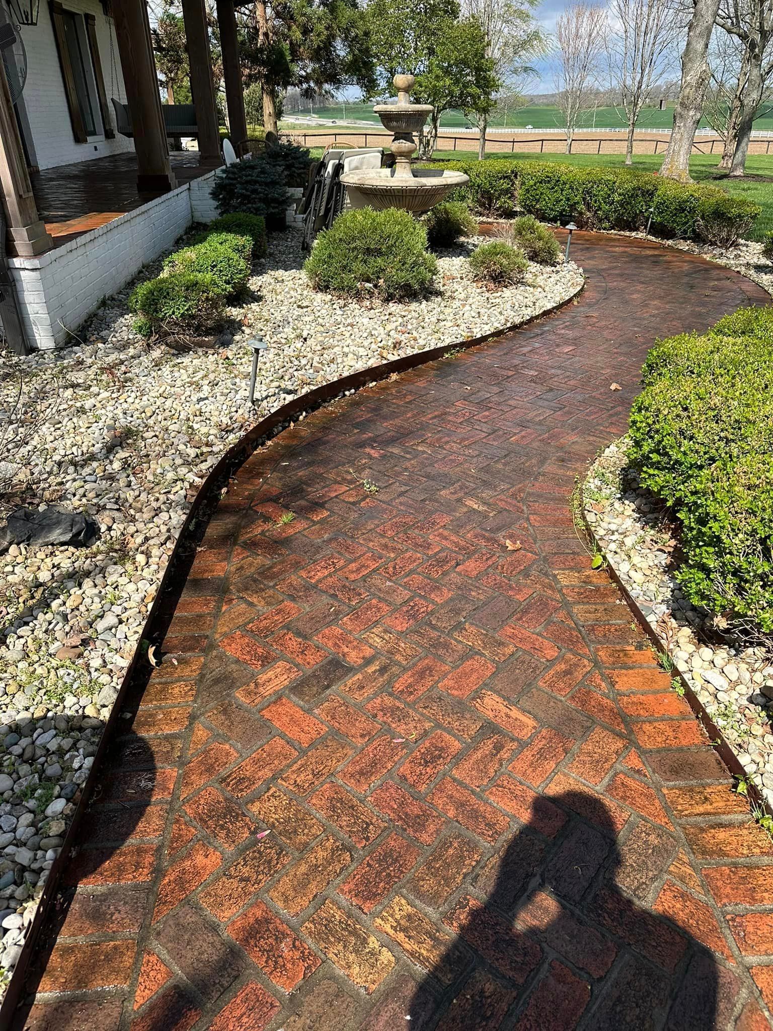 Brick pathway winds through a garden, bordered by rocks and green bushes, with a person's shadow in the foreground.