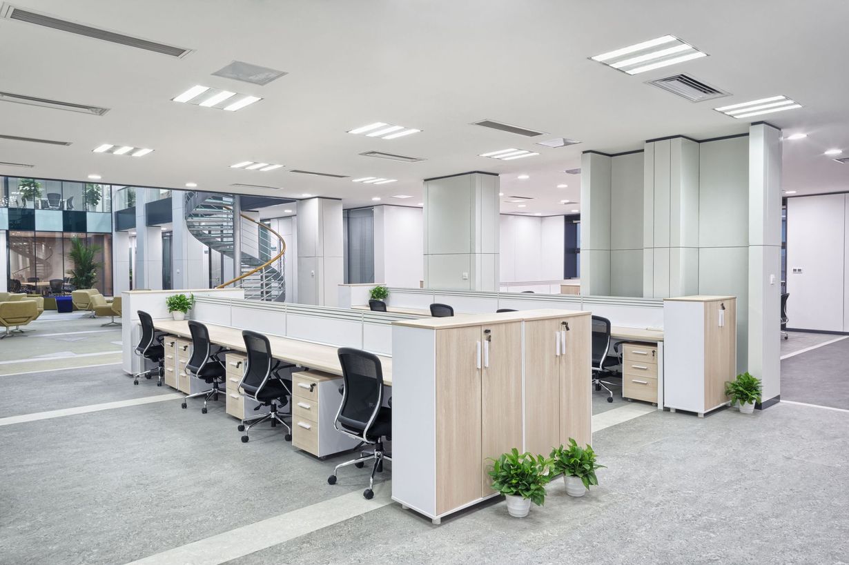 Modern office space with rows of desks, chairs, and storage cabinets. Bright overhead lighting and neutral colors.