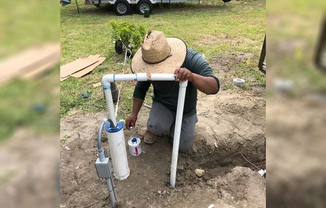 A man wearing a straw hat is kneeling down next to a pipe.