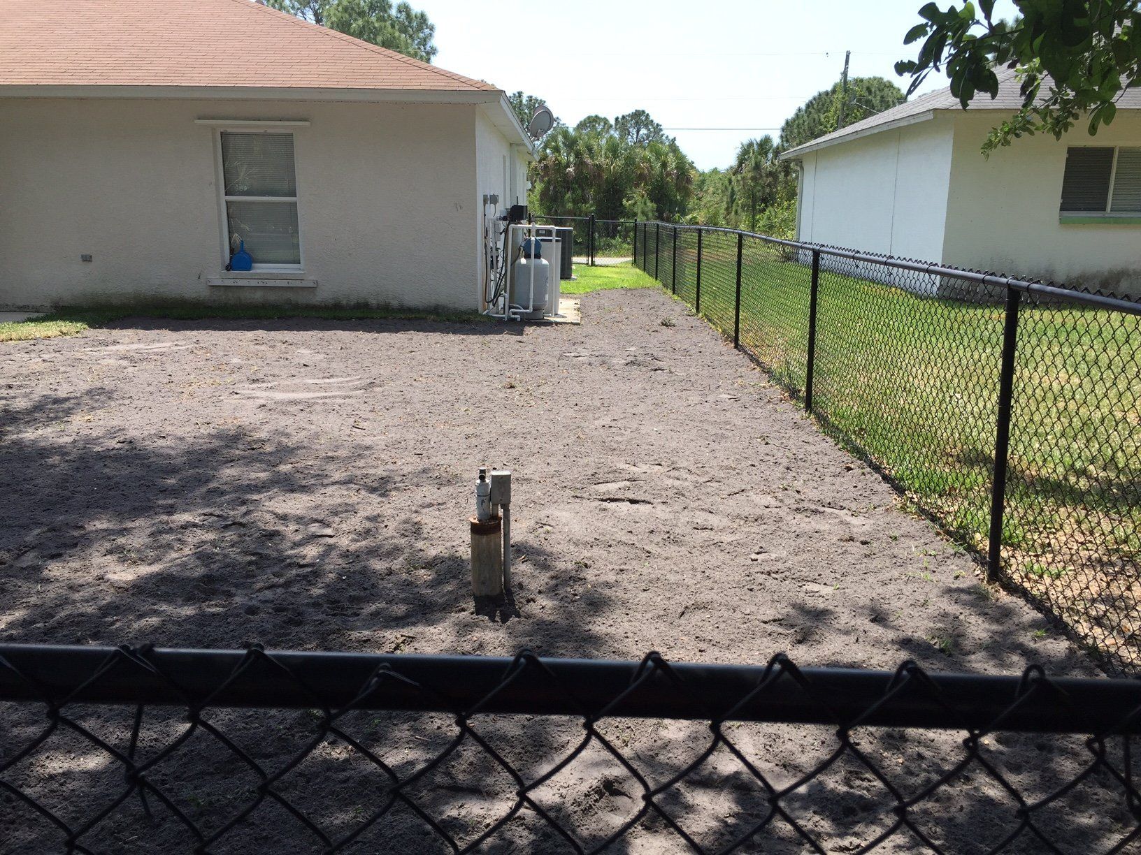 A chain link fence surrounds a dirt driveway leading to a house.