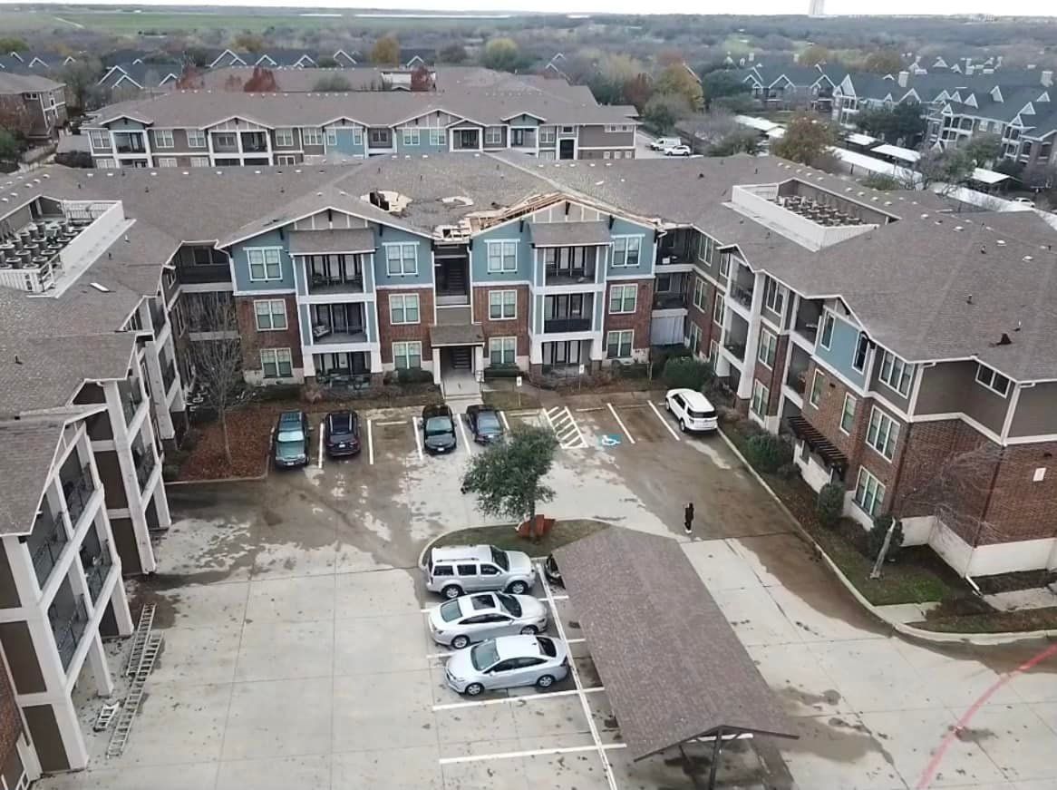 Aerial view of a multi-story apartment building with roof damage and parked cars. Overcast sky.