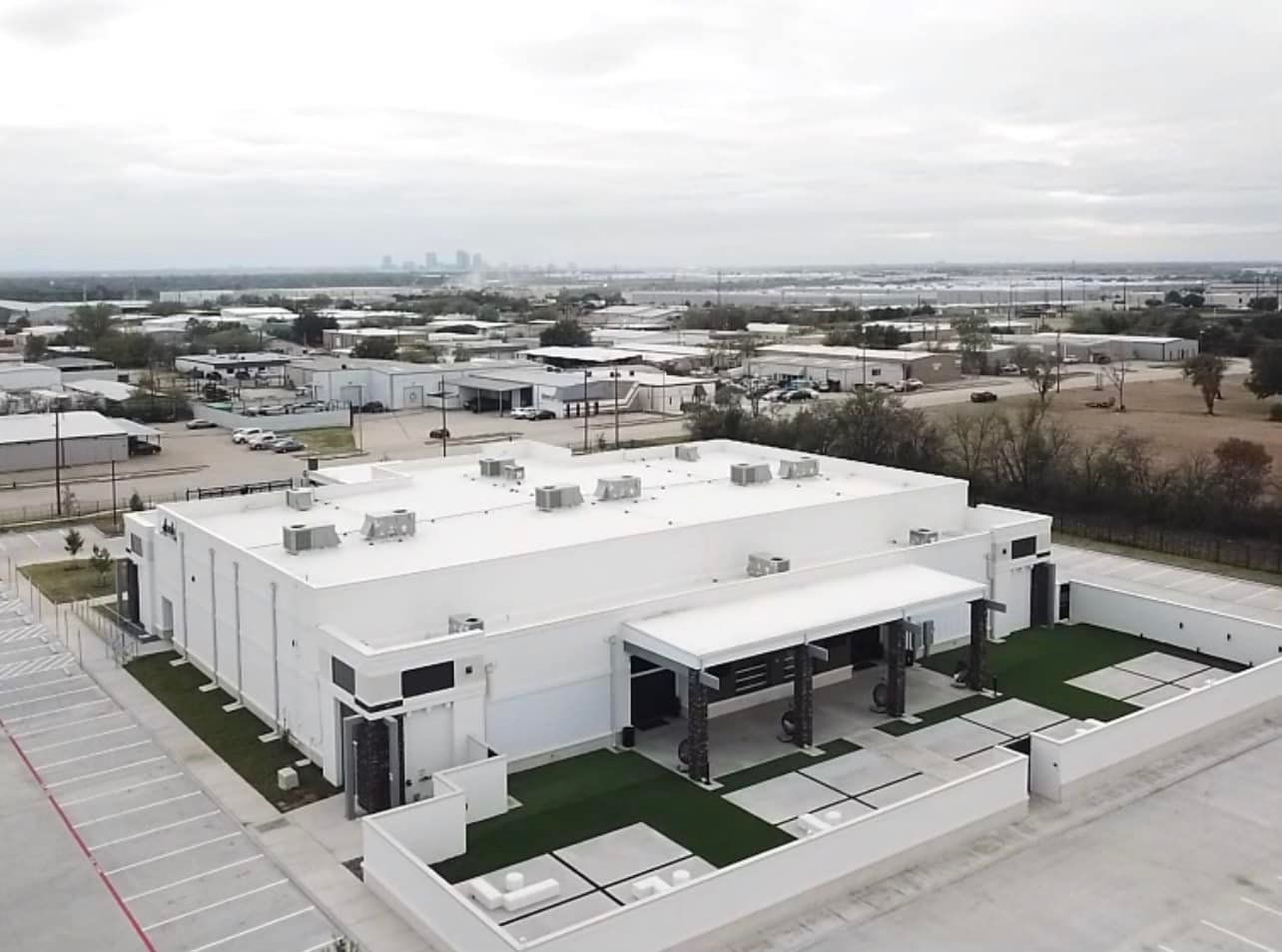 White industrial building with a flat roof, portico, and fenced-in lawn, with a city skyline in the distance.