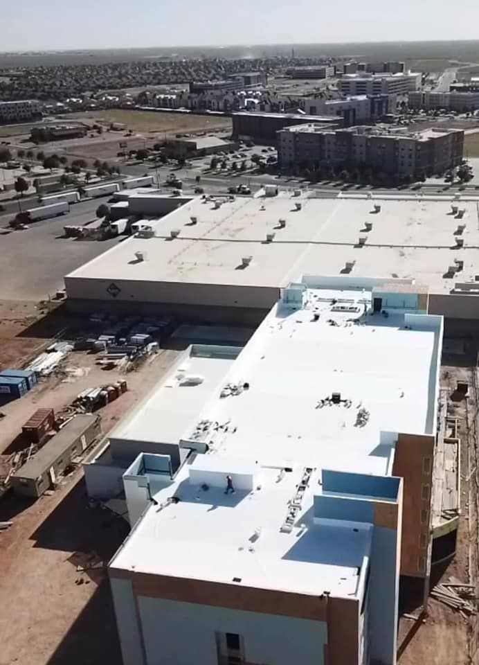 Aerial view of a building under construction, with a flat white roof. Nearby, a large shopping center and town in the background.