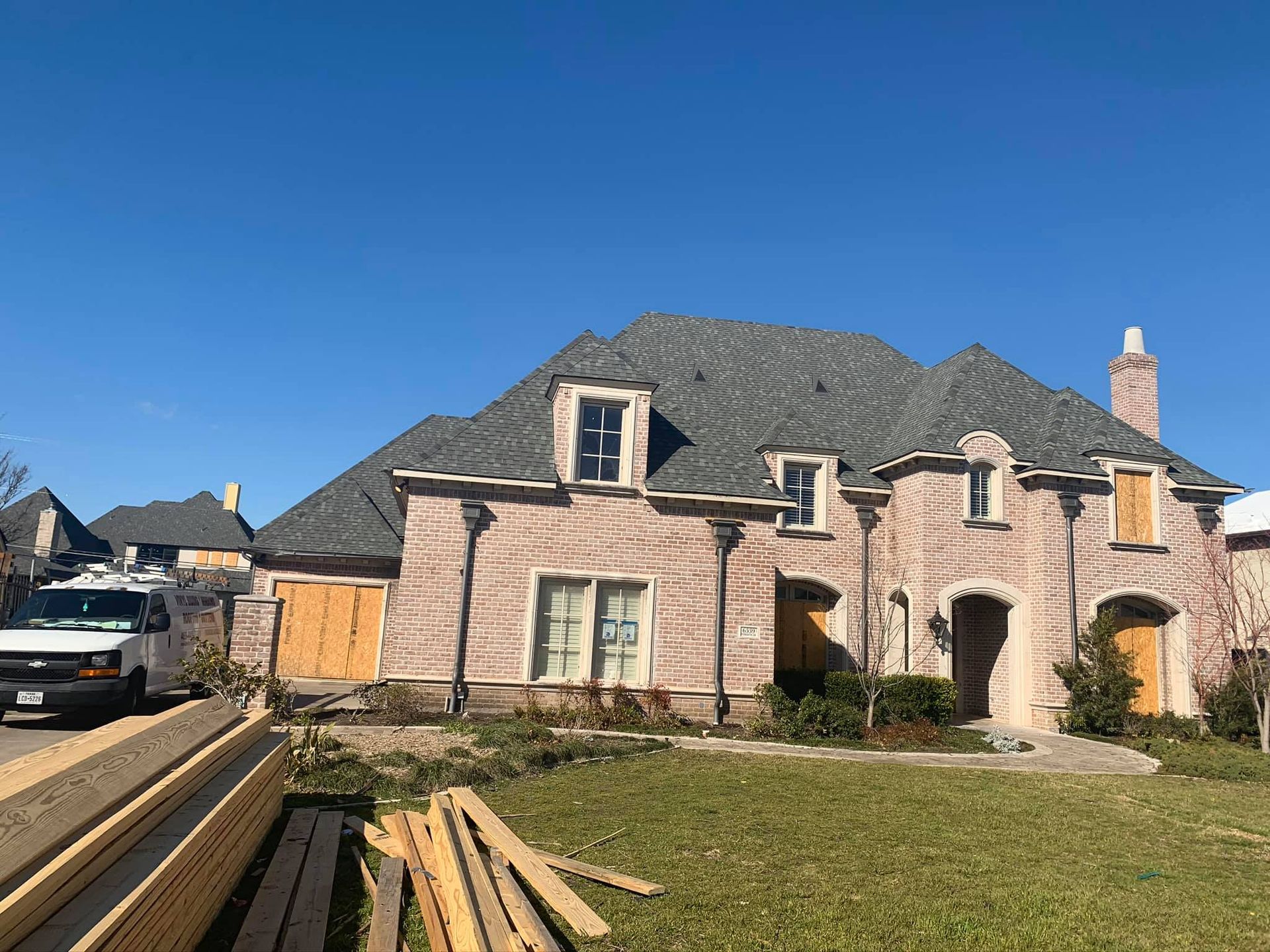 A brick house with a gray roof and boarded-up windows, construction materials in the foreground.