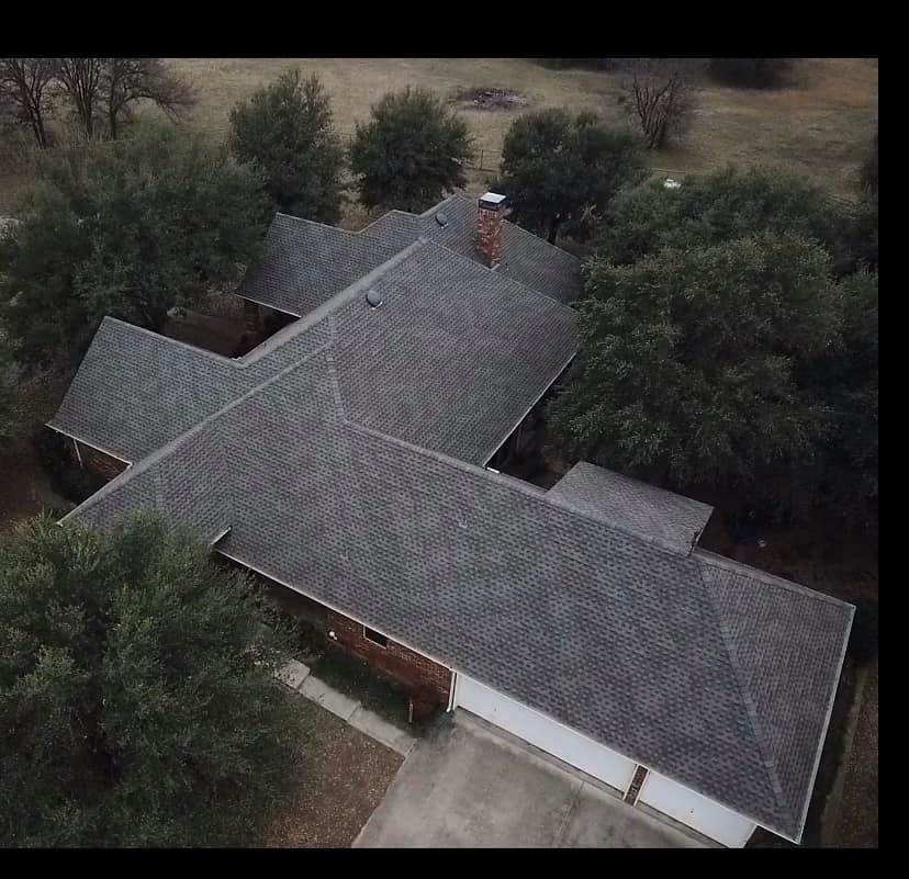 Aerial view of a house with a gray shingled roof, surrounded by trees. Includes a driveway and garage.