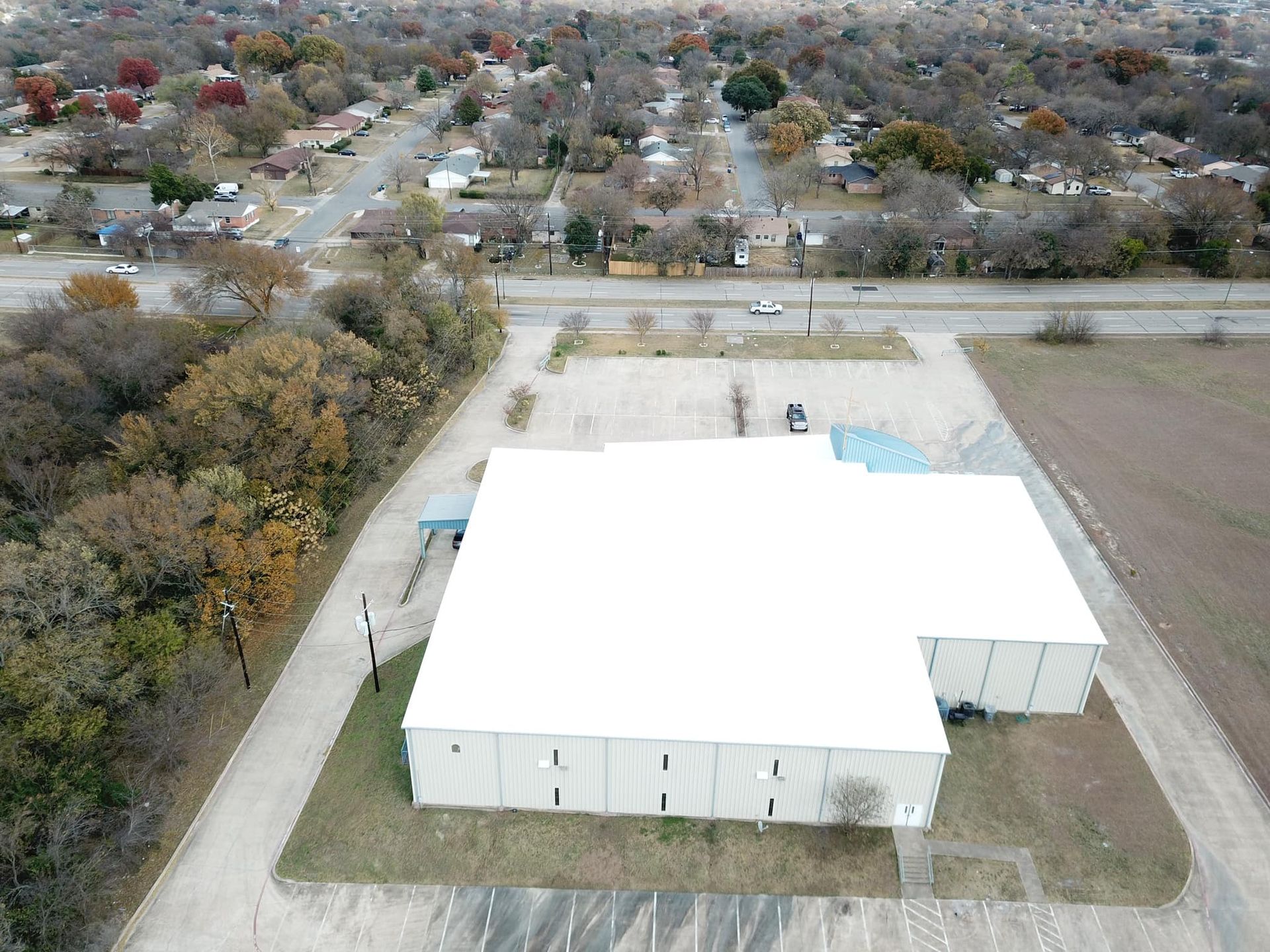 Aerial view of a large white building with a parking lot, near a road and neighborhood with autumn trees.