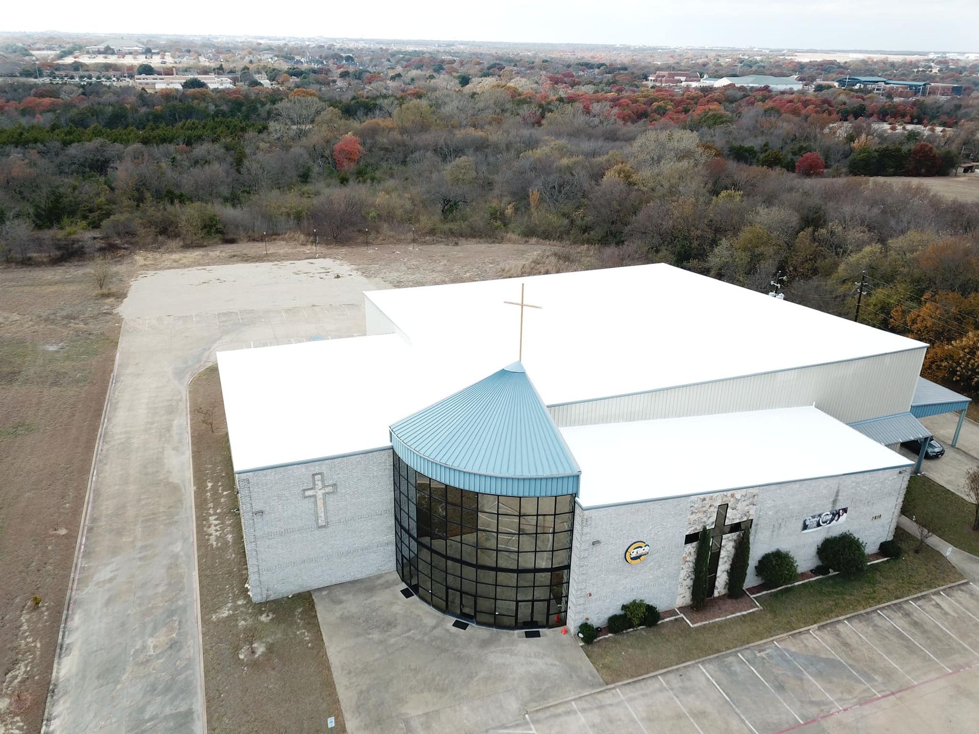 Church building with white roof and a blue-tiled section, a cross, and surrounding trees.
