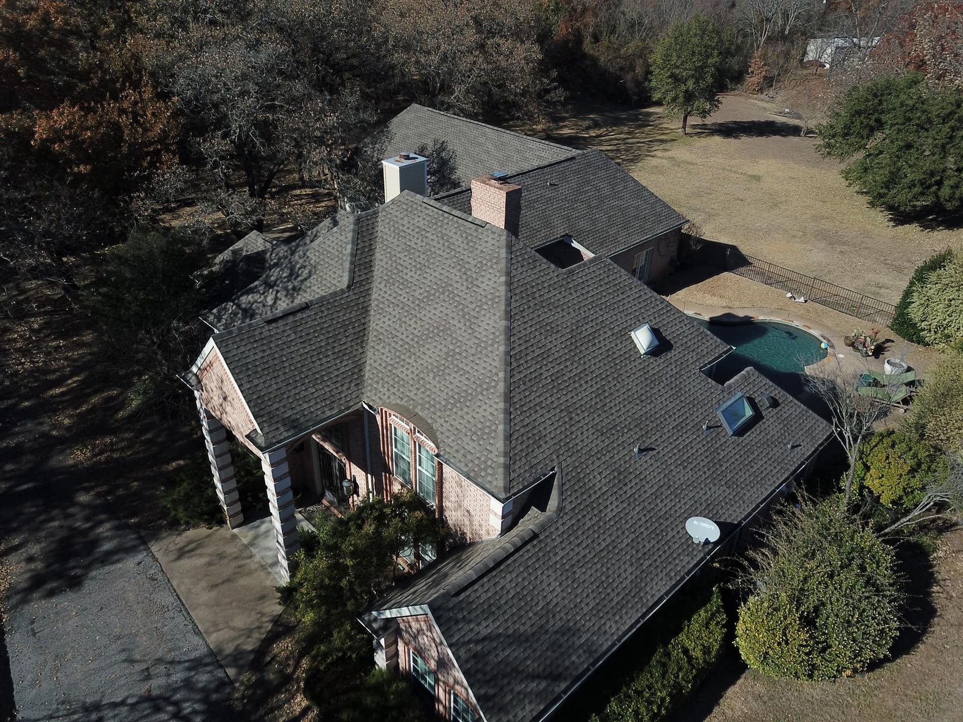 Aerial view of a house with a dark gray shingled roof, a chimney, and a pool in the backyard surrounded by trees.