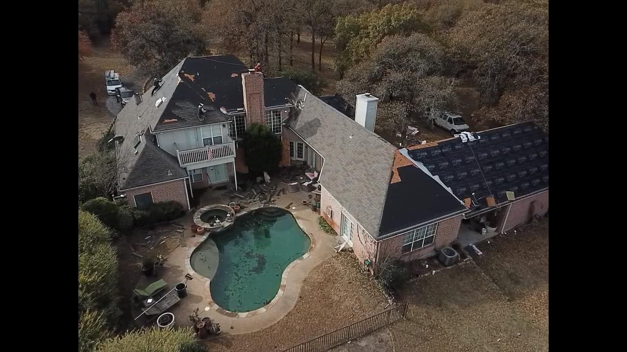 Aerial view of a house with a swimming pool and damaged roof. Surrounded by trees and grass.