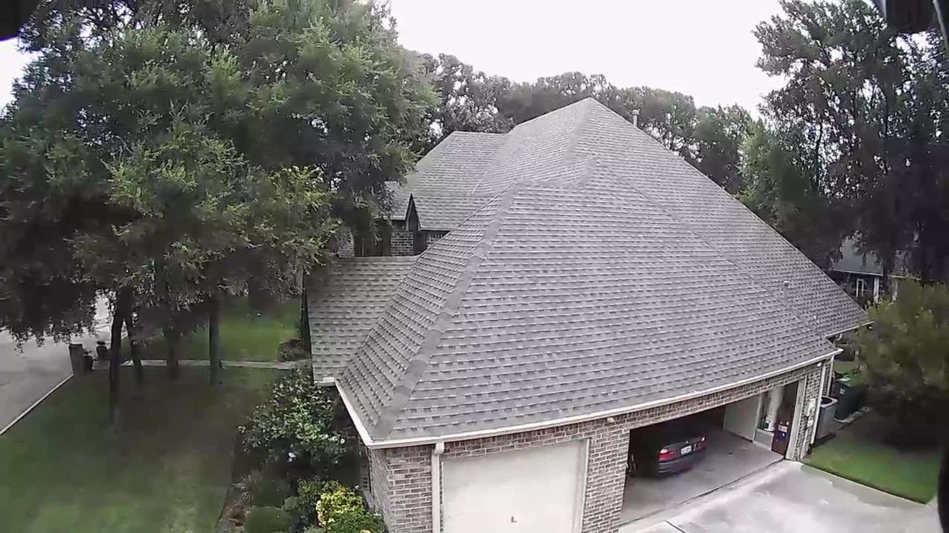 House with gray shingle roof, attached garage, and a car inside. Green trees and grass surround the house.