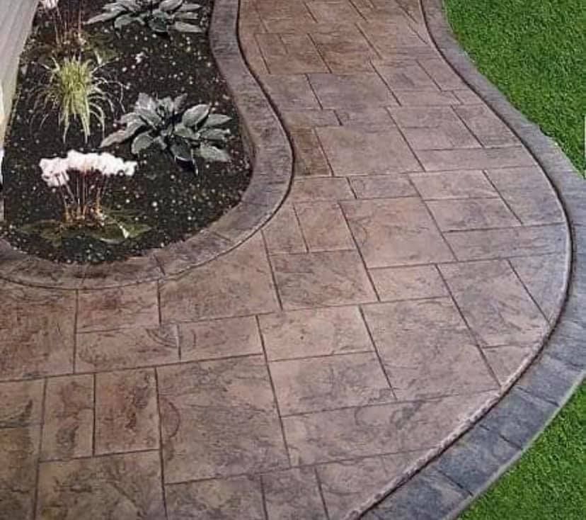 Stamped concrete pathway with flower bed. Brown paving with textured design, bordered by darker gray trim and green grass.