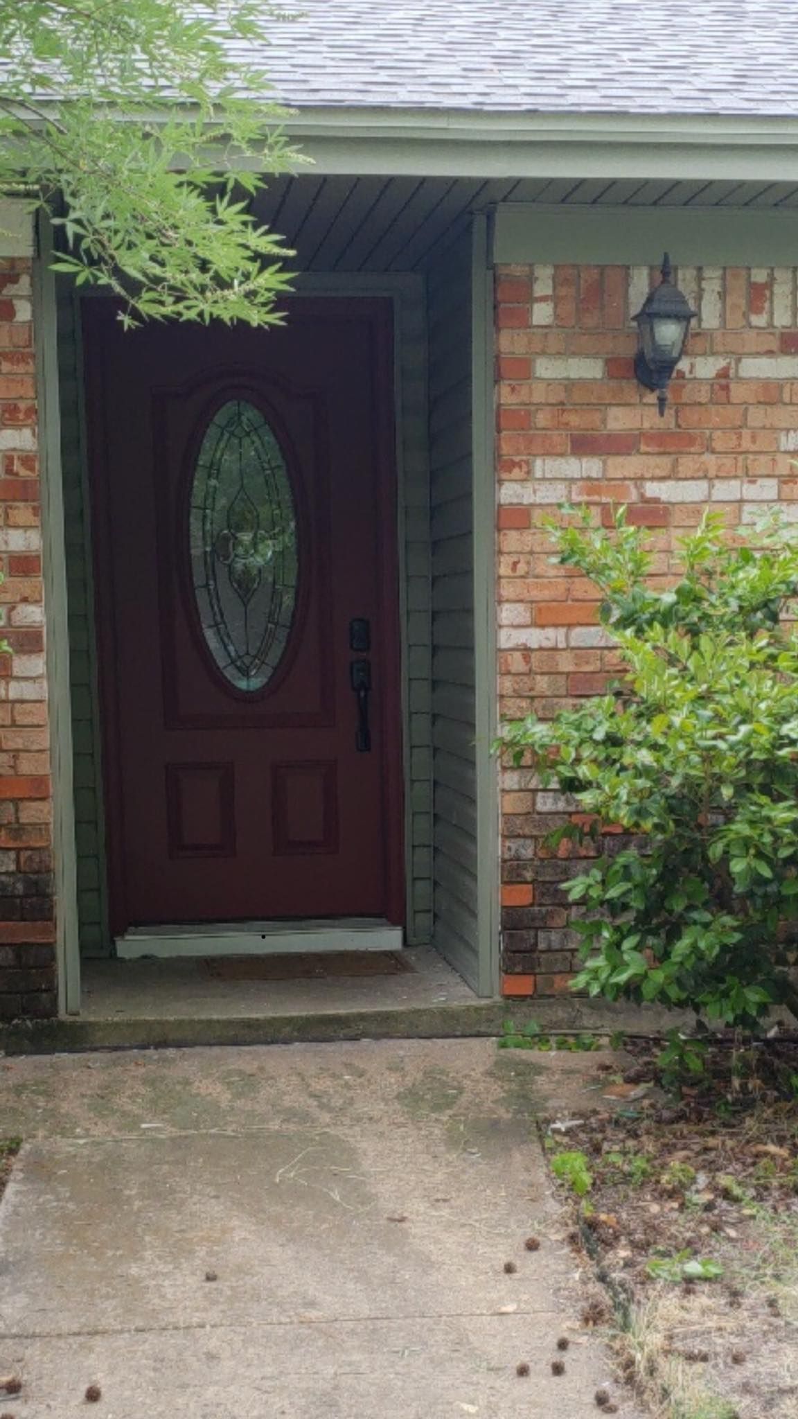 Red front door with oval window, brick exterior, concrete walkway, and bushes.