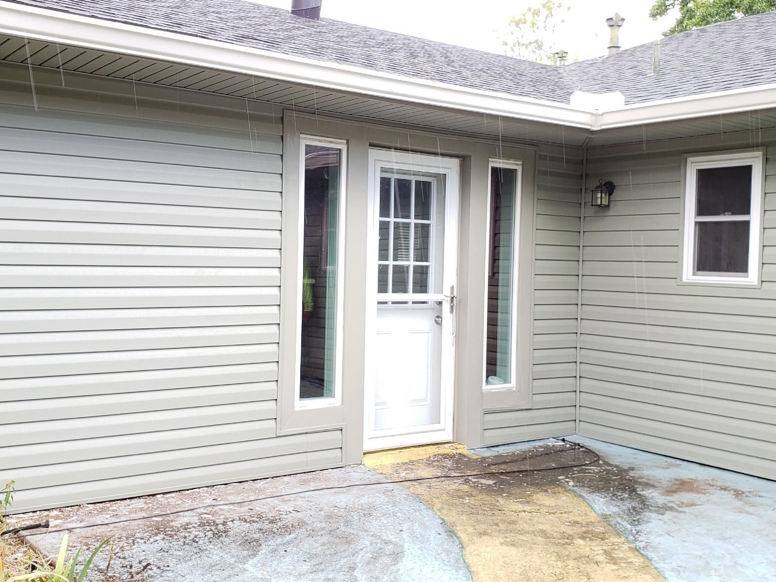 Exterior view of a house with a white door, two narrow windows, and gray siding. The ground shows water stains.