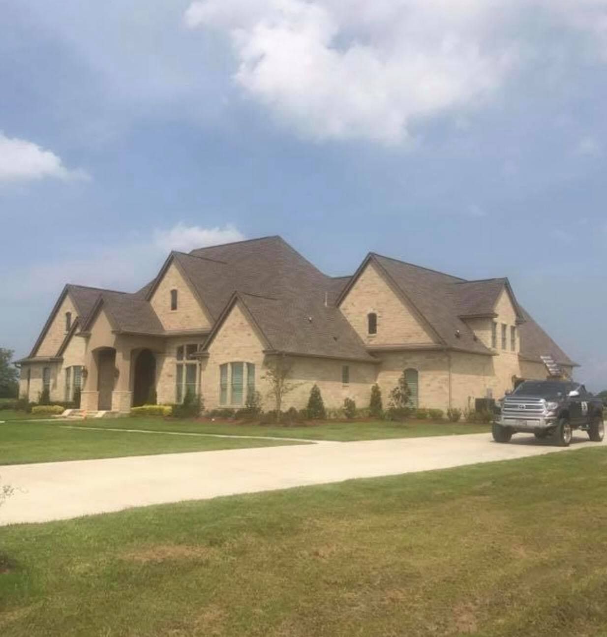 Large brick house with a brown roof and a truck in the driveway. Green lawn, blue sky with clouds.