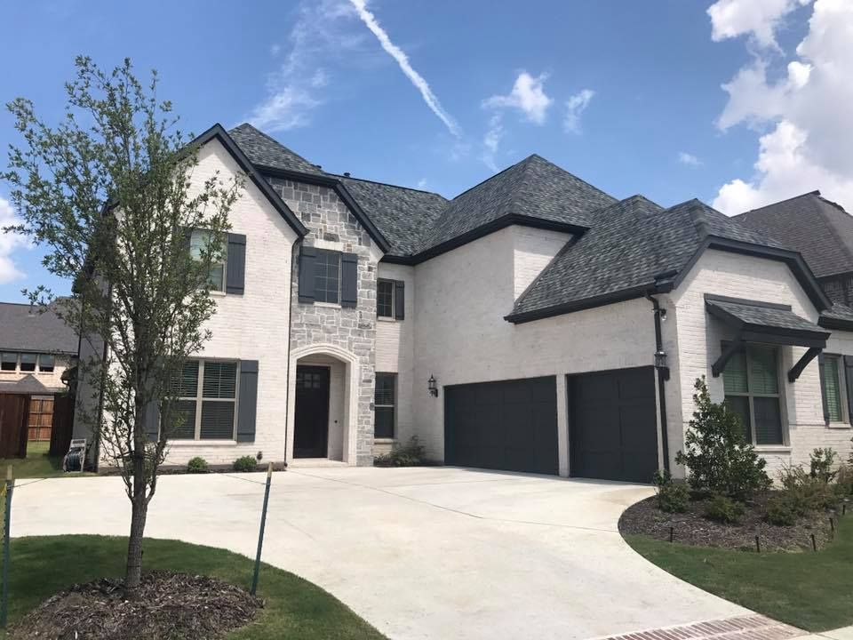 Two-story beige house with gray roof and garage doors, white trim, and a curved driveway on a sunny day.