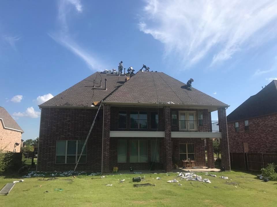Workers on a house roof replacing shingles on a sunny day. Brown brick house with green grass.